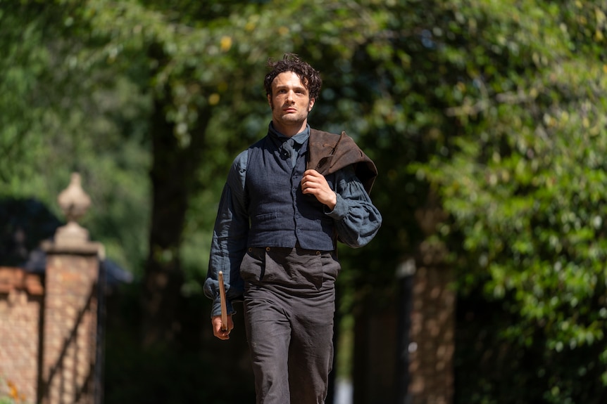 A man walks in his shirt and vest down a street with a determined expression, his coat thrown over his shoulder