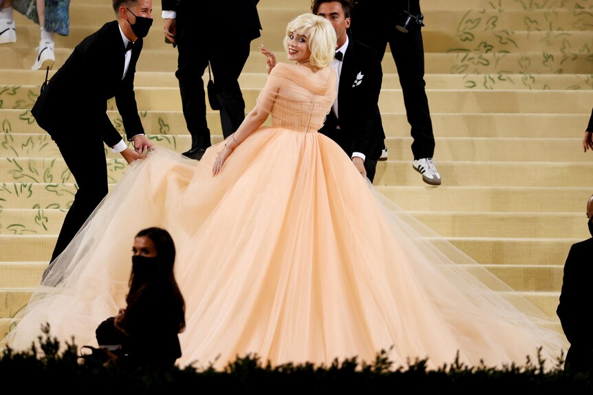 Billie Eilish looks back over her shoulder while wearing a peach tulle ball gown.