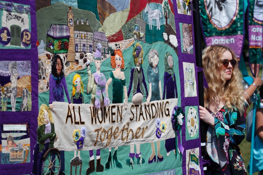 A woman holding a large banner showing women from different backgrounds and the words "all women standing together".