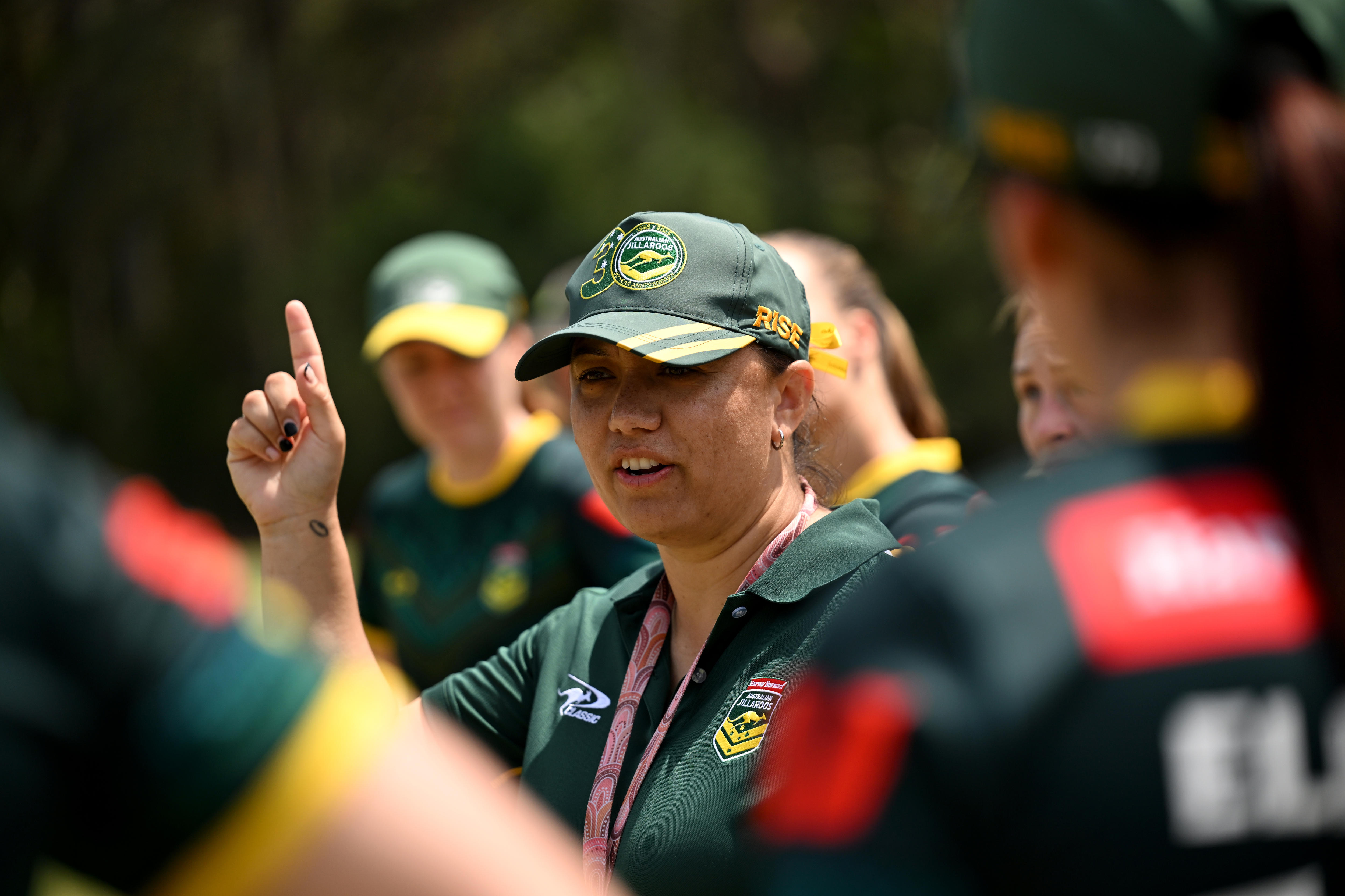 A woman in a green Jillaroos cap is pointing to the sky while talking to a circle of women around her.