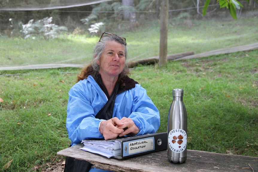 A woman in a blue jacket sits at an outdoor table with a large silver water bottle and folder.