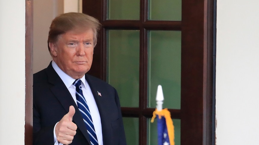 A man in a suit gives a thumbs up gesture with his right hand as he stands in the doorway of a building.