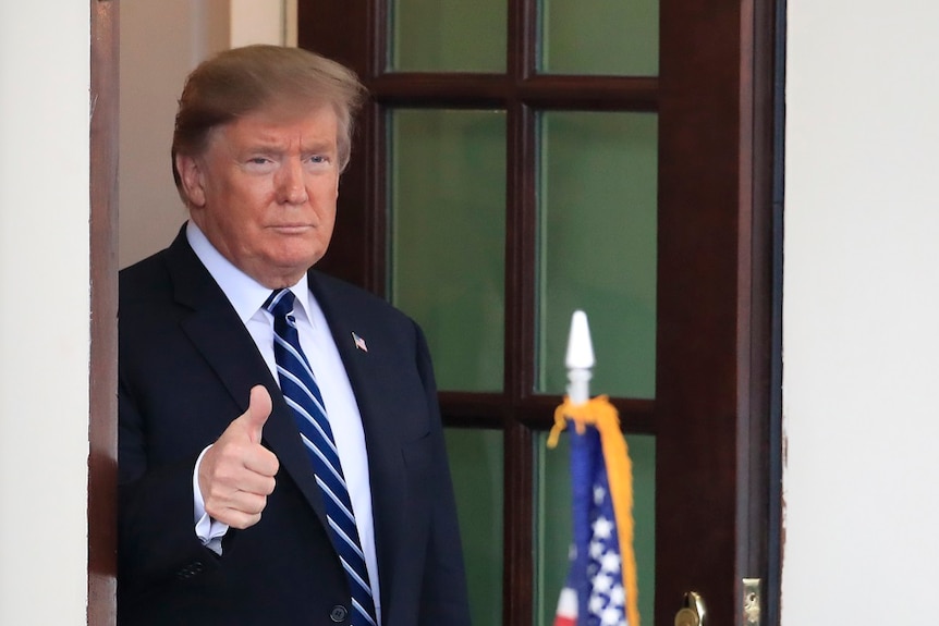 A man in a suit gives a thumbs up gesture with his right hand as he stands in the doorway of a building.