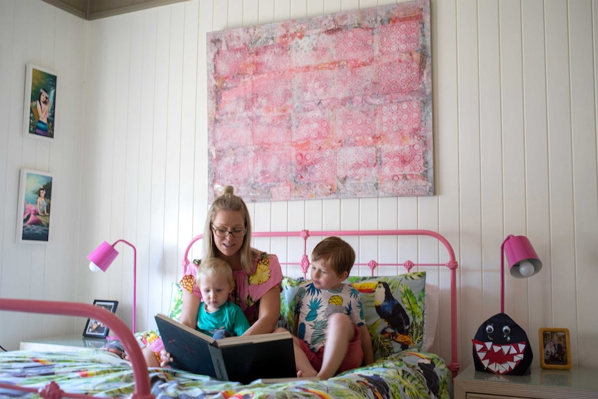 Jess Marsellos with her two sons, winston and Augustus, reads a story on a bed with artwork behind her.