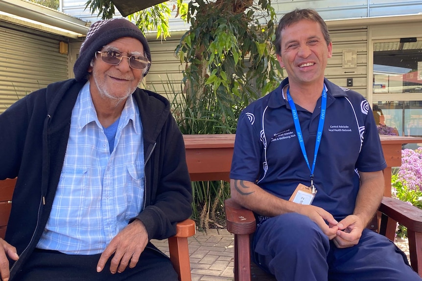 Two men sit on a bench under a tree smiling