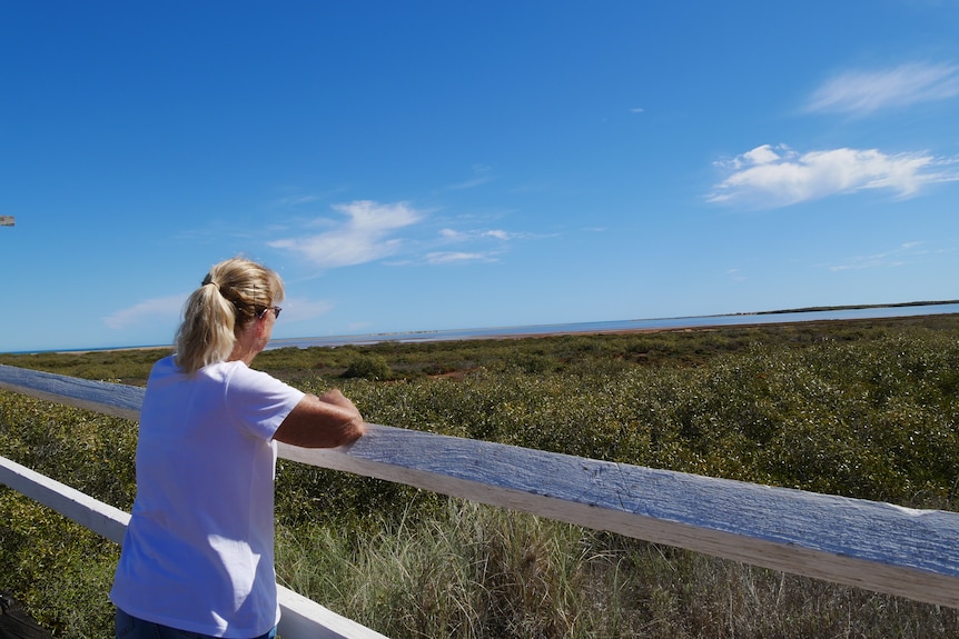 A woman in white tee, blonde hair tied back, leans against white wooden planks looking out to the mangroves and ocean. 