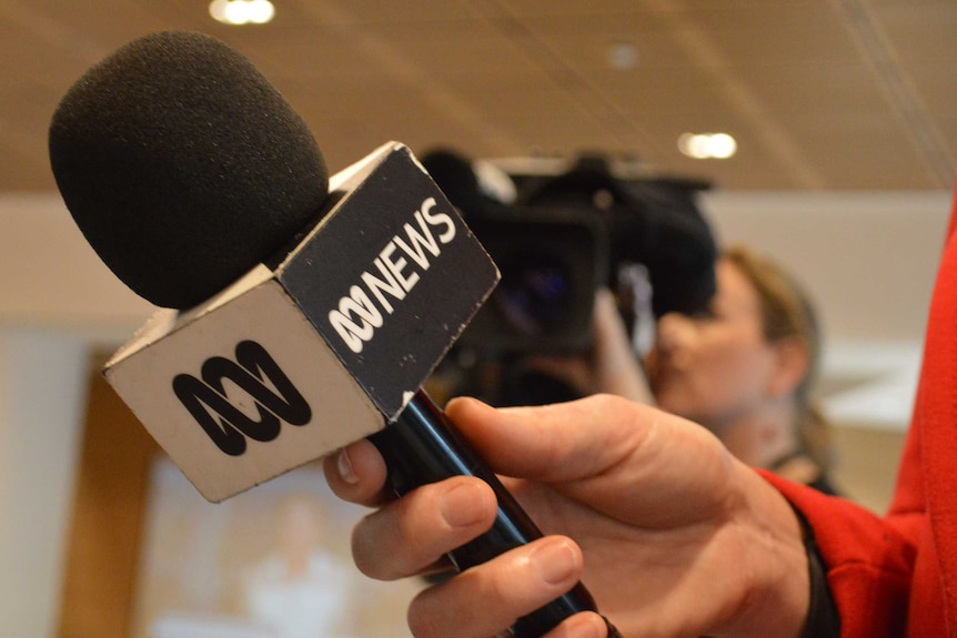 Close up of microphone with ABC News logo in hand with camera in the background.