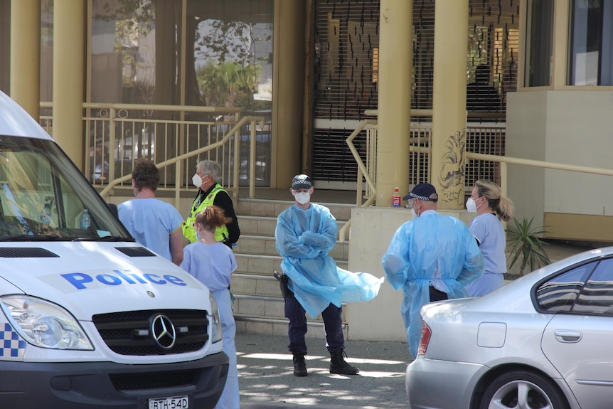 Six people standing in a street wearing personal protective equipment near police cars.