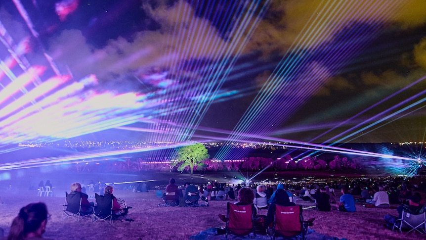 People sit on camping chairs watching a light show by a lake, including lasers, and a spot-lit tree