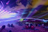 People sit on camping chairs watching a light show by a lake, including lasers, and a spot-lit tree