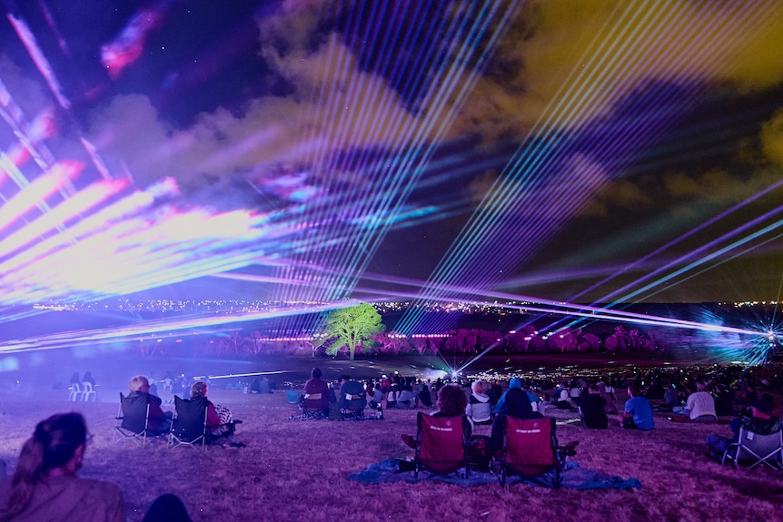 People sit on camping chairs watching a light show by a lake, including lasers, and a spot-lit tree