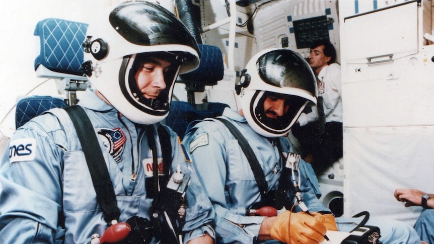 Two men in baby blue NASA jumpsuits are in a practice space shuttle with their helmet visors raised.