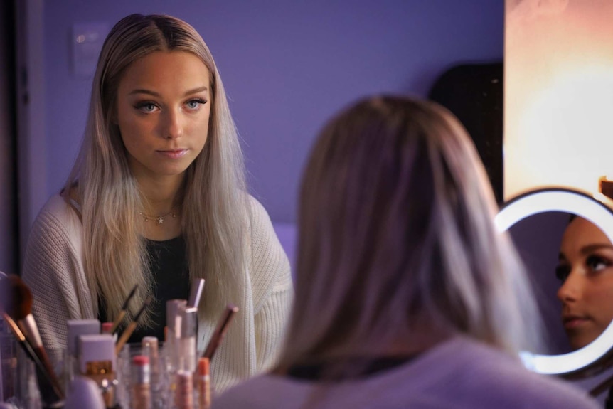 Olivia Plant looking into a mirror with long blonde hair and wearing a white cardigan, sitting at a table with make-up items