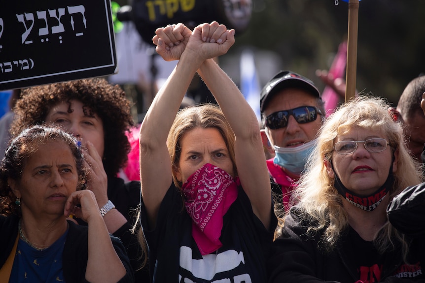 A blonde woman with red scarf over her face raises her arms above her head in a crowd of protesters.