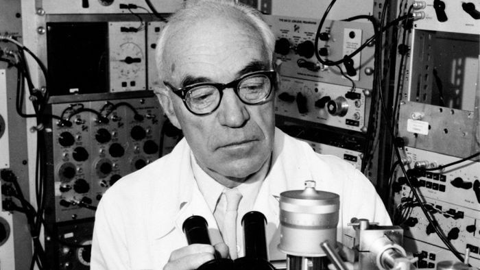 A man wearing thick glasses and a lab coat sits near a microscope and lots of science equipment.