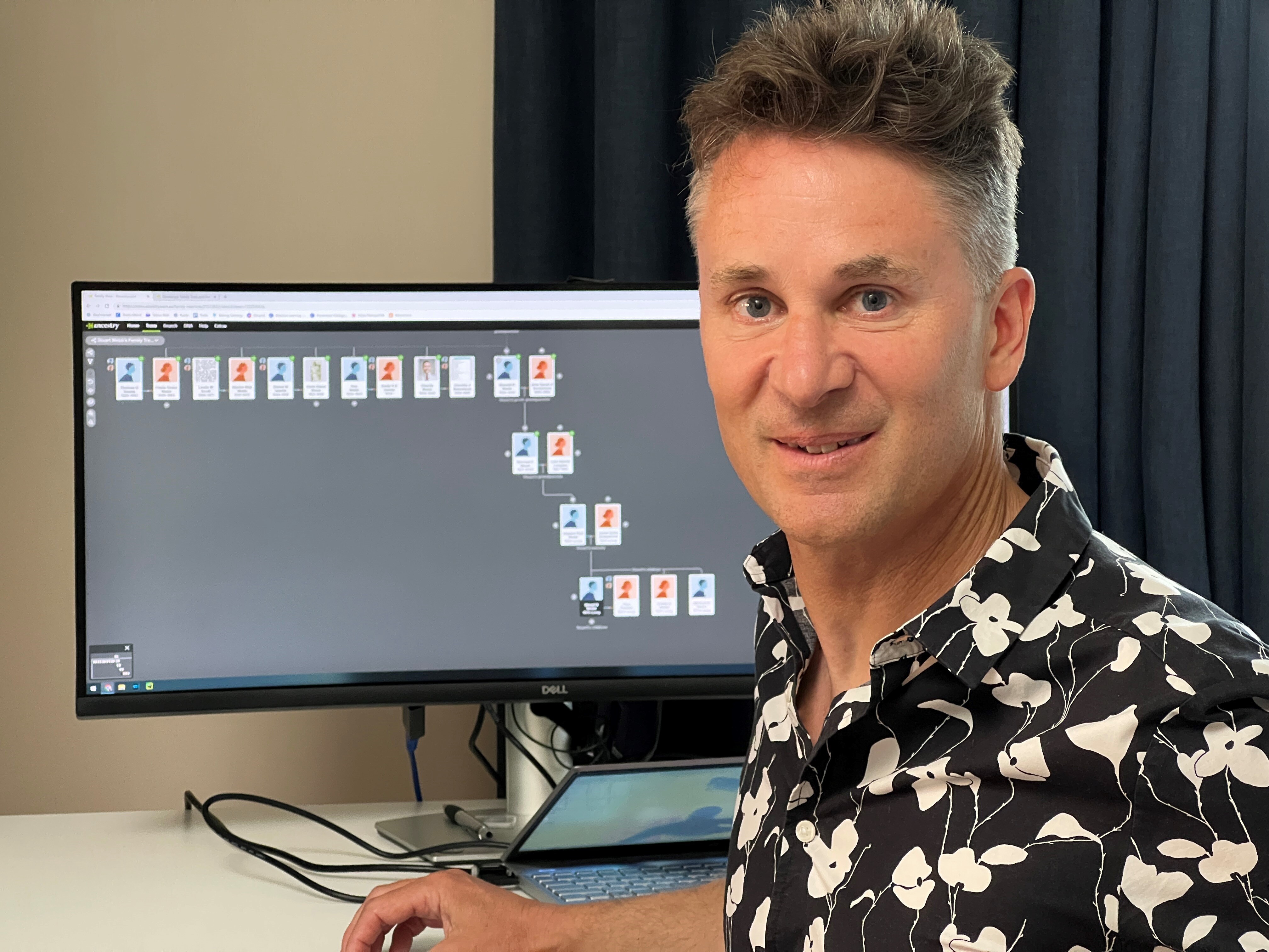 man wering a collared shirt sits in front of a computer showing a family tree