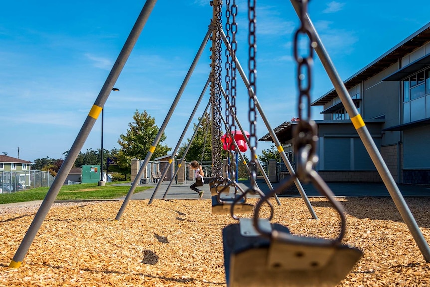A lone child sits on the last of a line of swings in a school playground.