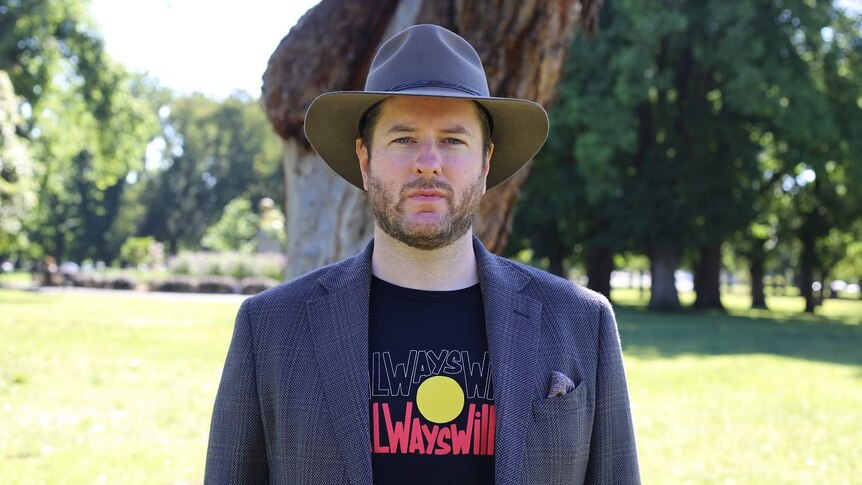 Marcus Stewart stands in a park, dressed in an Akubra and t-shirt bearing the message 'Always Was, Always Will Be'.