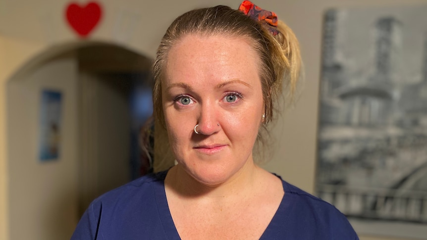 Kim Gallaher, dressed in her blue scrubs uniform, stands in her home.