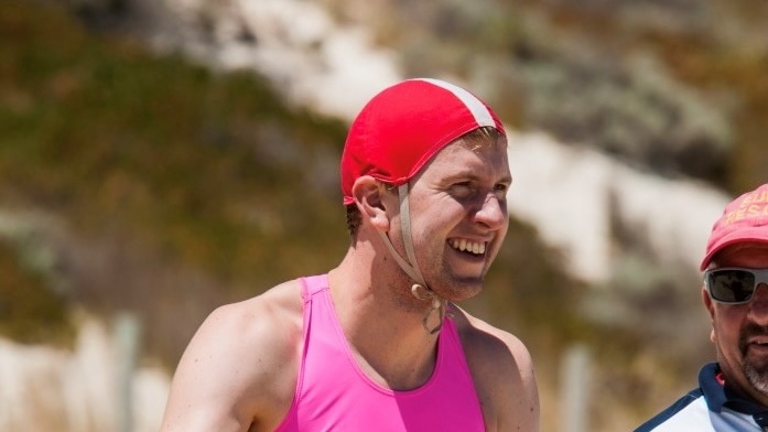 A smiling man in surf lifesaving gear prepares to enter the ocean with a paddle board under his arm