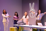 Catherine Văn-Davie sits on boardroom table with three young women standing around her, and neon hand sign for peace on wall.
