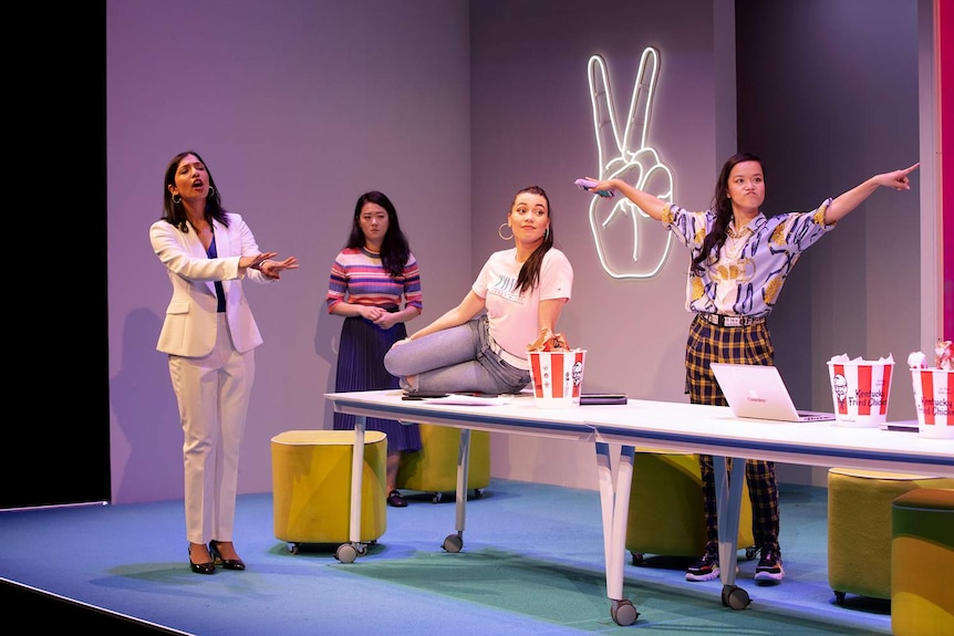 Catherine Văn-Davie sits on boardroom table with three young women standing around her, and neon hand sign for peace on wall.