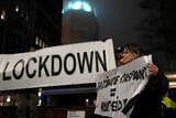 Woman holding a small  small anti-COVID restriction in front of a bigger sign with the word Lockdown written on it. 
