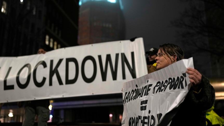 Woman holding a small  small anti-COVID restriction in front of a bigger sign with the word Lockdown written on it. 