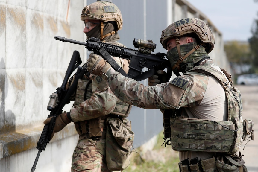 Two soldiers dressed in fatigues and helmets hold guns as they stare at a wall.