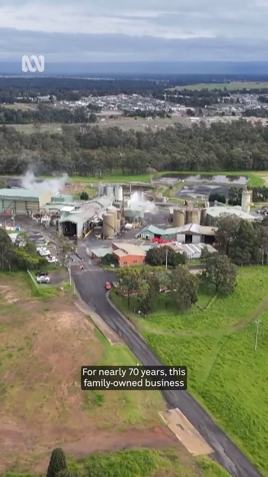 a drone shot shows an industrial premises surrounded by green paddocks with residential areas visible in the background