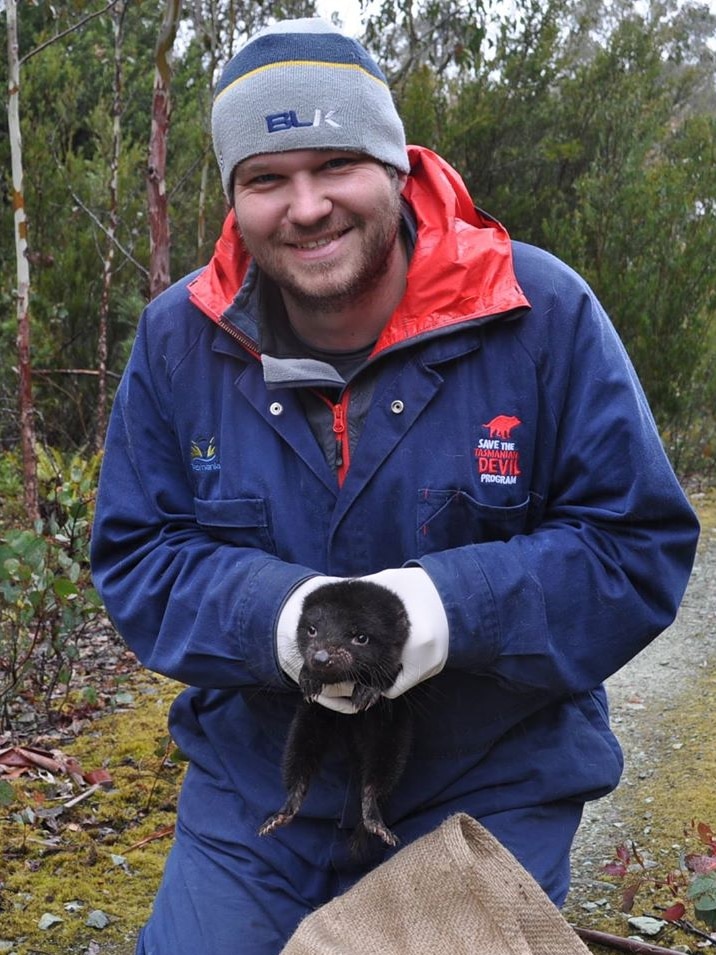 A man wearing a coat and beanie holds a young Tasmanian devil