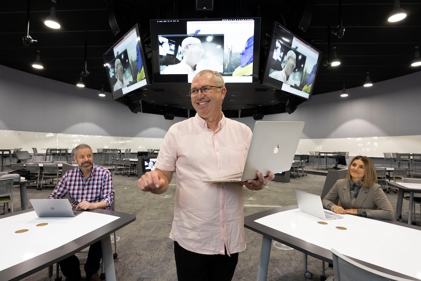 A man holding a laptop and smiling with two students sitting at desks in the background looking at him