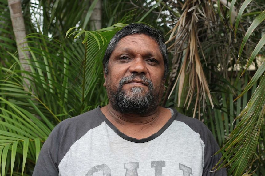 Indigenous man Lance Sullivan, standing in front of palm trees and wearing a t-shirt,  looks at the camera.