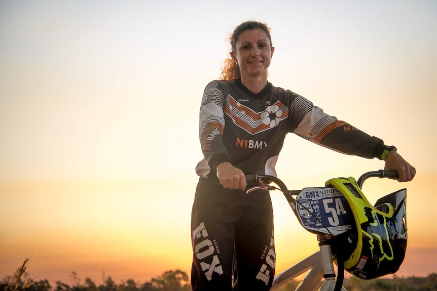 An older female BMX rider stands on the top of a dirt mound with her bike as the sun sets behind her.