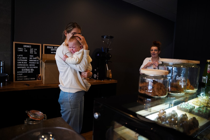 A young woman wearing jeans and a sweater behind a cafe till holds a small crying baby.