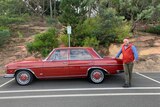 A man standing with a classic red Mercedes Benz in a car park with greenery behind him. 