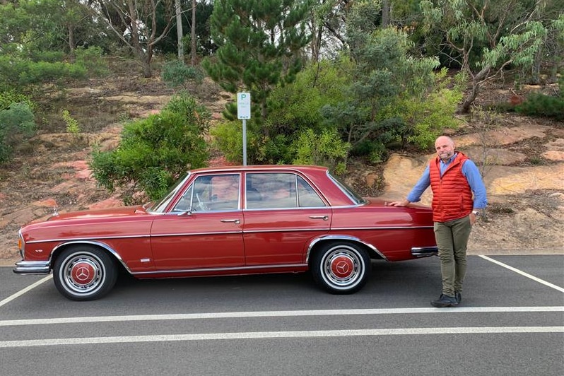 A man standing with a classic red Mercedes Benz in a car park with greenery behind him. 