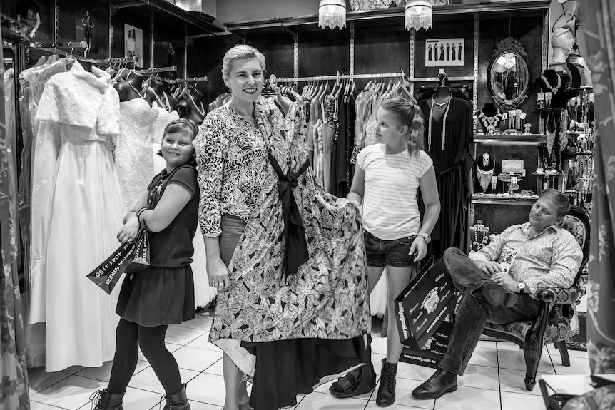 A woman and two young girls look through a clothing shop while a bored looking man sits on a chair in the corner.