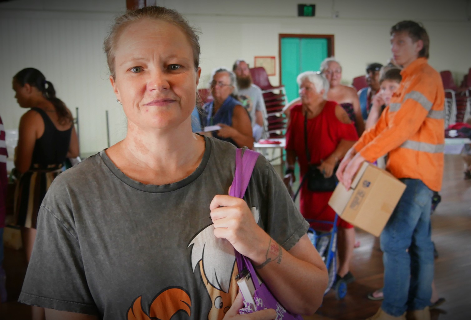 A woman holding a bag on her shoulder looking at the camera, in front of a queue of people