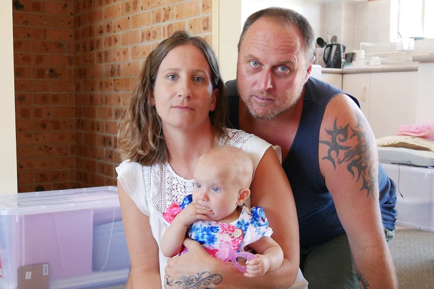 A woman and a man sit on the ground with a baby in a house, posing for a photo looking glum.