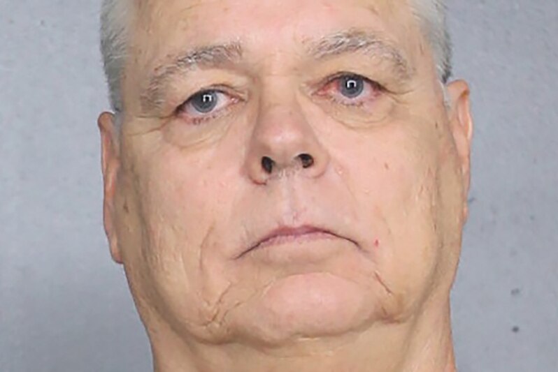 A clean shaven man with grey hair stares at the camera as his mugshot is taken.