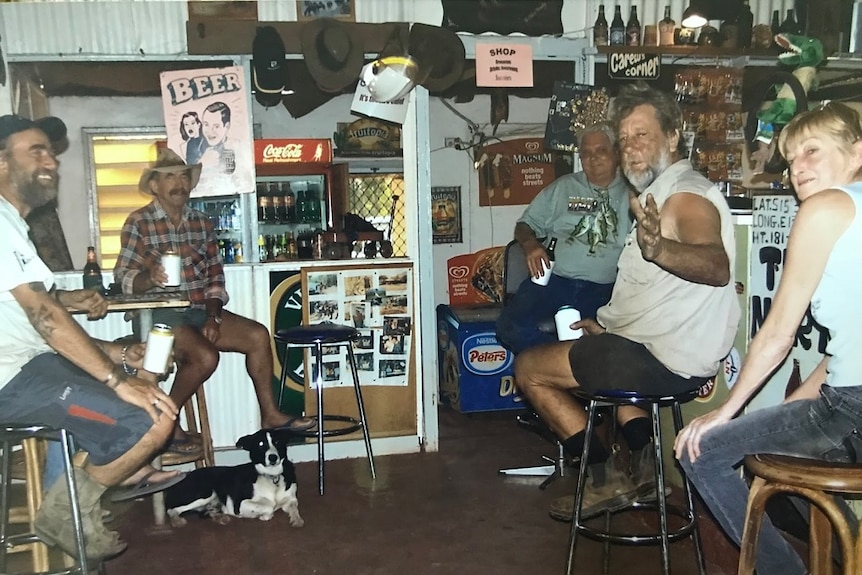 Five people sit around an outback pub drinking stubbies of beer held in coolers.