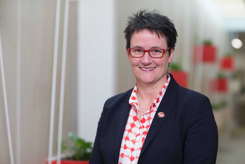 Meredith Peace, avec des lunettes rouges et un blazer noir, sourit à la caméra.