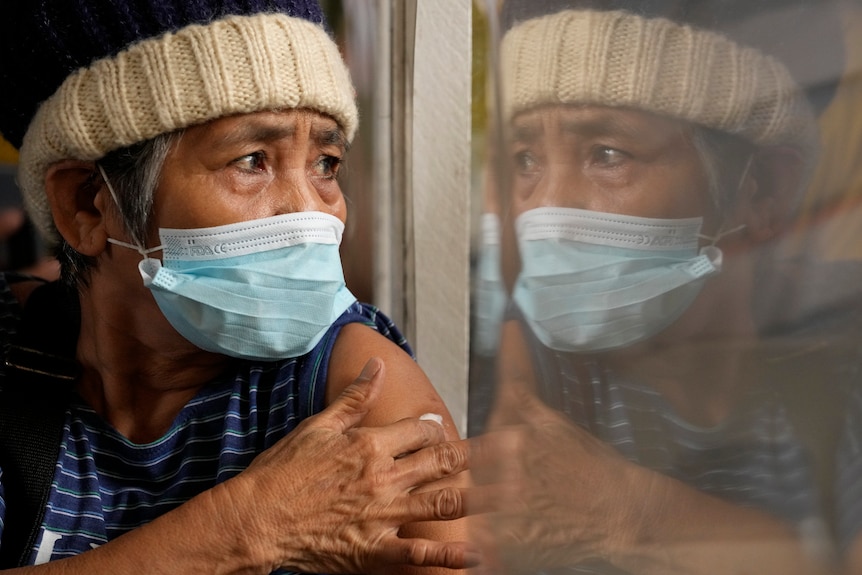 A woman in a face mask and a beanie clasps a cotton ball to her shoulder while gazing out the window