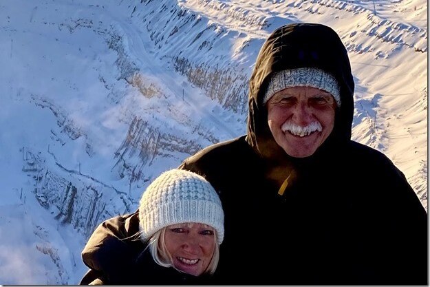 A couple in winter clothing with snow in the background smile