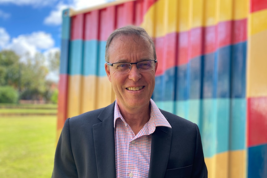 A man stands in front of colourful background smiling at the camera wearing a suit jacket and shirt 