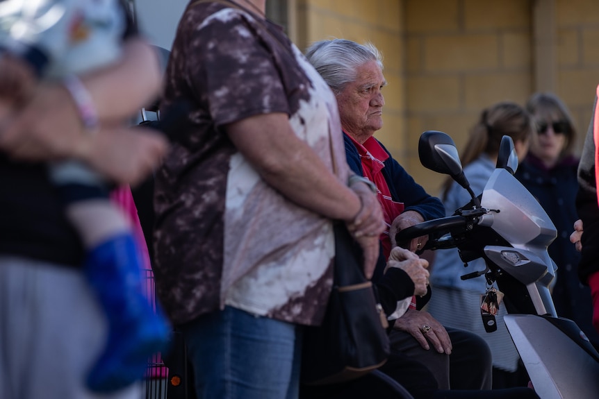 A person sits in a mobility scooter looking out at dozens of people gather outside a medical centre.