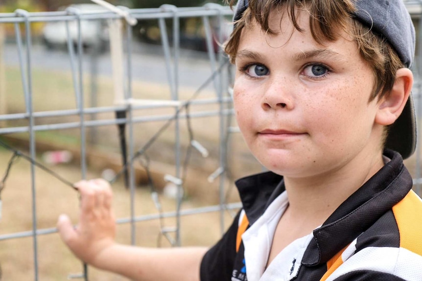 Primary school-aged boy standing beside a fence looking intently at camera