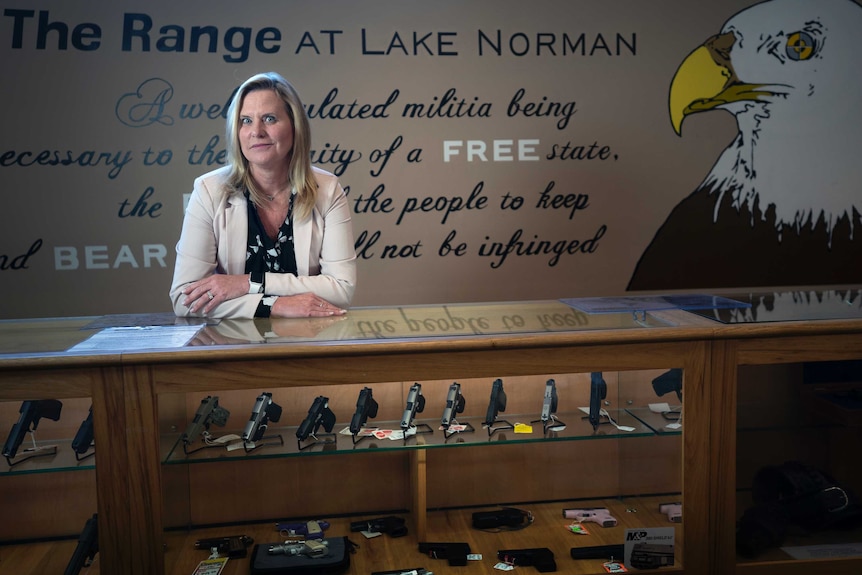 A woman in a cream blazer leans on a glass cabinet filled with guns