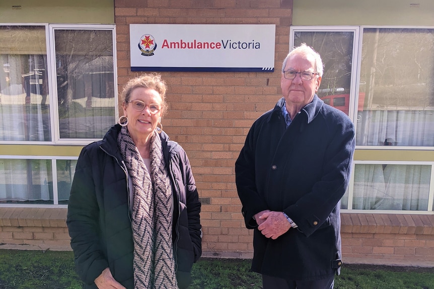 A man and woman, both wearing glasses and dark jackets, stand in front of a brick building with an Ambulance Victoria sign